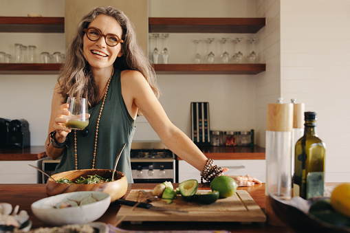 Healthy senior woman smiling while holding some green juice in her kitchen. Mature woman serving herself wholesome vegan food at home. Happy woman taking care of her aging body with a plant-based diet.